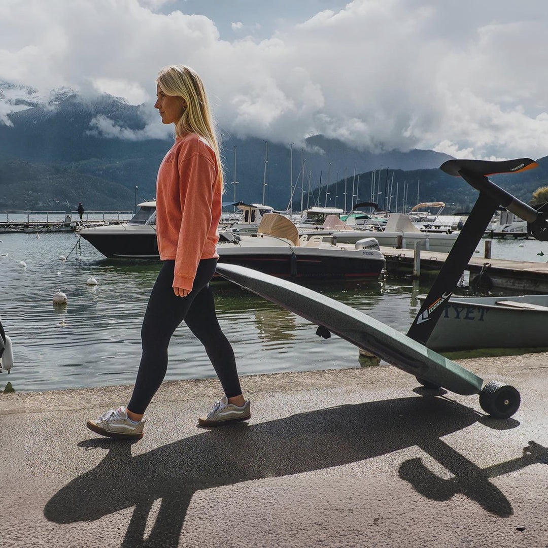 Woman walking with a long object near a dock with boats and mountains in the background